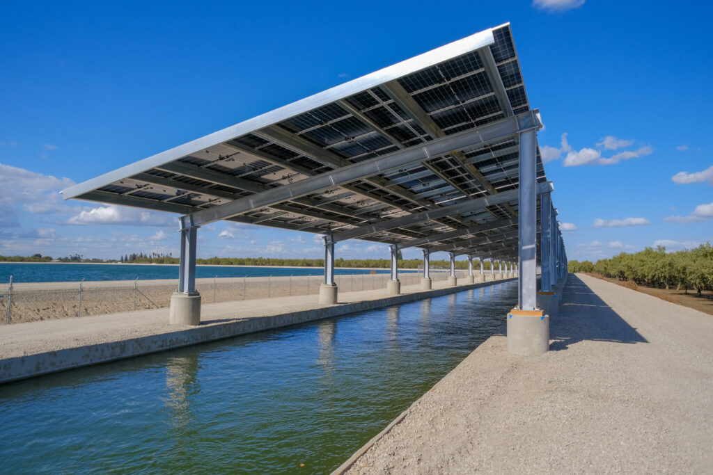 Under-panel view of a steel-truss solar canopy covering a Turlock Irrigation District canal on a clear day, showing shaded water and banks—Project Nexus, California.